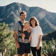 A young family — mum, dad and their baby — smiling together on a mountain hike