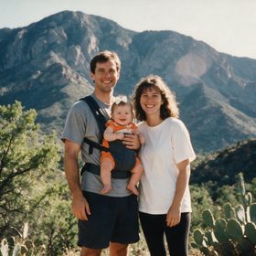 A young family — mum, dad and their baby — smiling together on a mountain hike